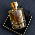 Maison Alhambra Karat by Maison Alhambra 100ml EDP — A clear glass perfume bottle with gold ornate detailing and a gold cap, resting on a black and gold square box, with a black tassel hanging from the neck. | Maison Alhambra Karat by Maison Alhambra 100ml EDP — Botol kaca jernih dengan perincian emas berornamen dan