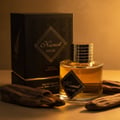Maison Alhambra Kismet Magic by Maison Alhambra 100ml EDP Unisex — A black textured box with gold geometric patterns and the Kismet Magic logo stands next to a clear glass perfume bottle filled with amber liquid, with two pieces of driftwood in the foreground and background. | Maison Alhambra Kismet Magic by Maison