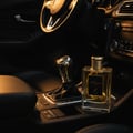 Maison Alhambra Kismet For Men by Maison Alhambra 100ml Man — A clear glass perfume bottle with a gold ribbed cap sits on a car's center console next to the gear stick, bathed in warm light. | Maison Alhambra Kismet For Men by Maison Alhambra 100ml Man — Botol kaca jernih dengan penutup berjalur emas terletak di