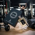 Maison Alhambra Kismet Moscow by Maison Alhambra 100ml EDP — A perfume bottle and its box are placed on a wooden surface with dumbbells and a towel in a gym setting. | Maison Alhambra Kismet Moscow by Maison Alhambra 100ml EDP — Botol minyak wangi dan kotaknya diletakkan di atas permukaan kayu dengan dumbbell dan
