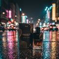 Maison Alhambra Exquisite Prive — A perfume bottle and its box sit on a wet, reflective street at night with blurred city lights in the background. | Maison Alhambra Exquisite Prive — Botol minyak wangi dan kotaknya diletakkan di jalan basah yang memantul pada waktu malam dengan lampu bandar yang kabur di latar