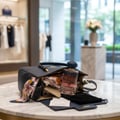 Maison Alhambra La Voie by Maison Alhambra 100ml for Her — A navy blue handbag is open on a marble table, with a bottle of La Voie perfume, a floral scarf, and a wallet inside. | Maison Alhambra La Voie by Maison Alhambra 100ml for Her — Beg beg tangan berwarna biru navy terbuka di atas meja marmar, dengan sebotol