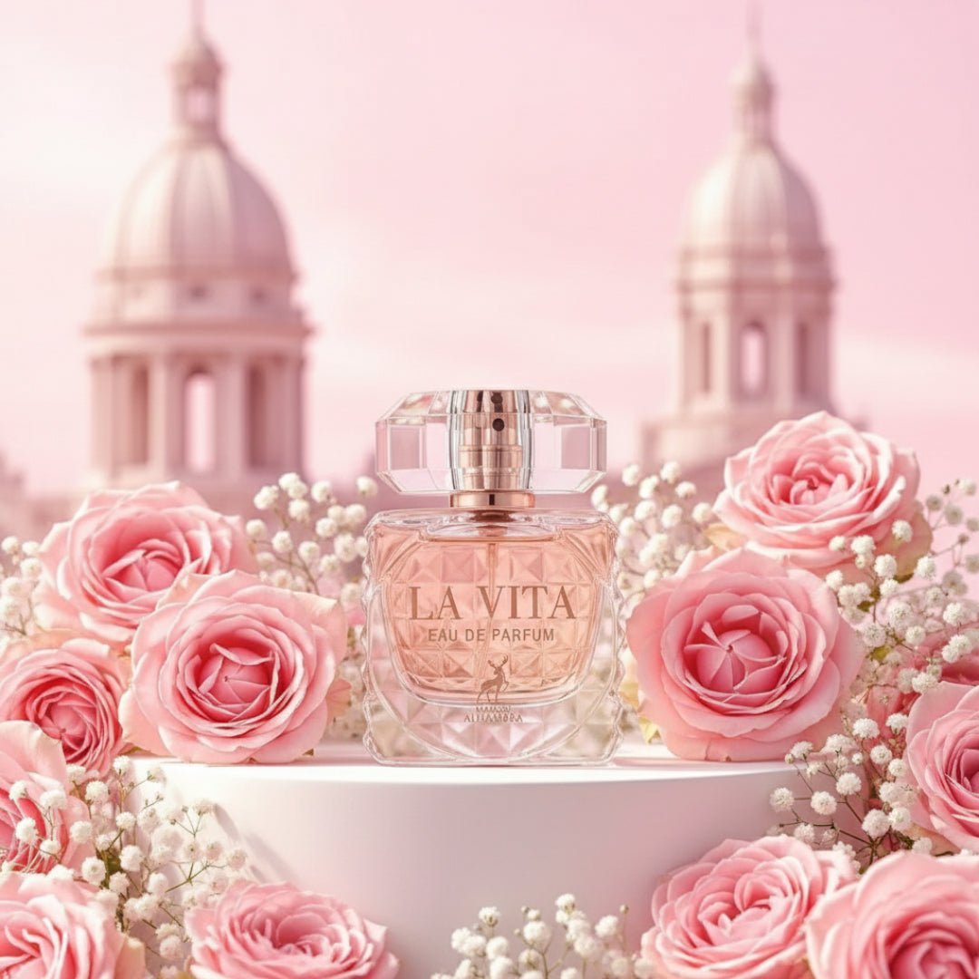 Maison Alhambra La Vita Maison Alhambra 100ml EDP Women — A faceted glass perfume bottle with a rose gold cap sits on a white pedestal, surrounded by pink roses and baby's breath, with blurred pink domed buildings in the background. | Maison Alhambra La Vita Maison Alhambra 100ml EDP Women — Botol minyak wangi kaca