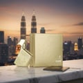 Maison Alhambra Luxe Gold – A gold perfume bottle and its box sit on a marble surface with a blurred city skyline and twin towers in the background. A passport and boarding pass are also visible. | Maison Alhambra Luxe Gold – Botol minyak wangi emas dan kotaknya diletakkan di atas permukaan marmar dengan latar