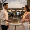 Maison Alhambra Luxe Gold by Maison Alhambra – 100 ml Unisex — A man and a woman smiling at each other across a marble table displaying a gold perfume box and bottle, with bokeh lights in the background. | Maison Alhambra Luxe Gold by Maison Alhambra – 100 ml Unisex — Seorang lelaki dan wanita tersenyum antara satu