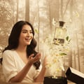 Maison Alhambra Avant by Maison Alhambra 100ml EDP Men — A smiling woman in a white dress reaches out her hands as a perfume bottle explodes with flowers and powder | Maison Alhambra Avant by Maison Alhambra 100ml EDP Men — Wanita tersenyum berbaju putih menghulurkan tangan ketika botol minyak wangi meletup dengan