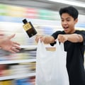 Maison Alhambra B.A.D. Homme by Maison Alhambra 100 ml for Men — A young man in a black t-shirt joyfully catches a flying perfume bottle while holding two white plastic shopping bags. | Maison Alhambra B.A.D. Homme by Maison Alhambra 100 ml for Men — Seorang lelaki muda berbaju hitam dengan gembira menangkap botol