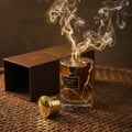 Maison Alhambra Exclusif Tabac — A clear glass perfume bottle with golden cap and brown liquid, emitting wisps of smoke, sits next to its open brown box on a woven mat. | Maison Alhambra Exclusif Tabac — Botol minyak wangi kaca jernih dengan penutup keemasan dan cecair coklat, mengeluarkan kepulan asap, terletak di