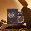 Maison Alhambra Infini Elixir — A purple perfume bottle and its box are displayed on a rocky surface with sand and pebbles, featuring gold geometric patterns. | Maison Alhambra Infini Elixir — Botol minyak wangi ungu dan kotaknya dipamerkan di permukaan berbatu dengan pasir dan kerikil, menampilkan corak geometri emas.