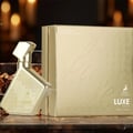 Maison Alhambra Luxe Gold – Gold perfume bottle and box with embossed circular pattern and text, set against a softly blurred background of warm lights. | Maison Alhambra Luxe Gold – Botol dan kotak minyak wangi emas dengan corak bulat timbul dan teks, berlatar belakang cahaya hangat yang kabur.
