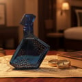 Maison Alhambra Luxe Journey by Maison Alhambra – 100 ml Unisex — A blue glass perfume bottle with gold geometric patterns sits on an old map, next to two small compasses and a thick book. The background is a dimly lit hotel room with a bed and lamp. | Maison Alhambra Luxe Journey by Maison Alhambra – 100 ml Unisex —
