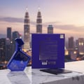 Maison Alhambra Luxe Nightlife Unisex 100 ml — A blue, geometric perfume bottle with silver speckles sits on a marble surface in front of a blurred cityscape at dusk, with a blue gift box and passport nearby. | Maison Alhambra Luxe Nightlife Unisex 100 ml — Botol minyak wangi biru geometrik dengan bintik perak