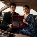 Maison Alhambra Luxe Street by Maison Alhambra – 100 ml Unisex — A man in a black suit and a woman in a sequined blue dress sit in a car, holding an open burgundy gift box containing a black perfume bottle and a black rectangular item. | Maison Alhambra Luxe Street by Maison Alhambra – 100 ml Unisex — Seorang lelaki