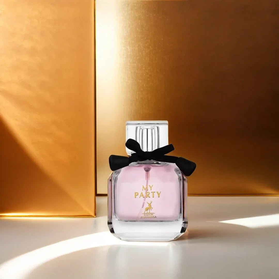 Maison Alhambra My Party Maison Alhambra 100ml EDP Woman — A clear glass perfume bottle with pink liquid and a black bow around the neck, set against a shimmering gold background with soft light casting shadows. | Maison Alhambra My Party Maison Alhambra 100ml EDP Woman — Botol kaca jernih berisi cecair merah jambu