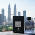 Maison Alhambra Maitre De Blue by Maison Alhambra 100ml for Men — A black box and a clear glass perfume bottle with blue liquid are placed on a surface with a cityscape and skyscrapers in the background. | Maison Alhambra Maitre De Blue by Maison Alhambra 100ml for Men — Kotak hitam dan botol minyak wangi kaca jernih