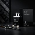 Maison Alhambra Man Black Edition — Black perfume bottle and box with silver accents, next to a watch and cufflinks on a dark textured surface, with a patterned fabric in the background. | Maison Alhambra Man Black Edition — Botol dan kotak pewangi hitam dengan aksen perak, bersebelahan jam tangan dan manset di