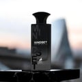 Maison Alhambra Mindset Eau de Parfum — A black perfume bottle and its white and black patterned box are displayed against a white background. The box features bold black lettering for 'MINDSET' and a deer logo. | Maison Alhambra Mindset Eau de Parfum — Botol minyak wangi hitam dan kotaknya yang bercorak hitam putih