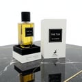 Maison Alhambra The Tux by Maison Alhambra 90ml EDP for Men — A clear glass perfume bottle with yellow liquid and a black cap, displayed on a black and white stand next to its white product box on a marble surface. | Maison Alhambra The Tux by Maison Alhambra 90ml EDP for Men — Botol minyak wangi kaca jernih dengan
