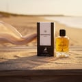 Maison Alhambra Montaigne Coco — A perfume bottle and its box sit on a sandy surface with a sheer fabric blowing in the background on a beach at sunset. | Maison Alhambra Montaigne Coco — Sebotol minyak wangi dan kotaknya diletakkan di atas permukaan berpasir dengan fabrik jarang tertiup angin di latar belakang di