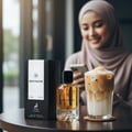 Maison Alhambra Montaigne Coco Maison Alhambra 100ml EDP — A perfume bottle and its box sit on a table next to a tall glass of iced coffee with whipped cream and toppings, with a woman in a hijab smiling at her phone in the blurred background. | Maison Alhambra Montaigne Coco Maison Alhambra 100ml EDP — Sebotol minyak
