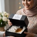 Maison Alhambra Montaigne Coco Maison Alhambra 100ml EDP — A smiling woman in a hijab and embellished dress opens a black gift box revealing a glass perfume bottle nestled in white shredded paper. | Maison Alhambra Montaigne Coco Maison Alhambra 100ml EDP — Seorang wanita tersenyum memakai hijab dan gaun berhias