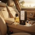 Maison Alhambra Montaigne Vanille by Maison Alhambra 100ml — A clear glass perfume bottle with a gold cap and a deer emblem sits on a car's center console next to its black and white box, with plush leather seats and a palm tree-lined cityscape in the background. | Maison Alhambra Montaigne Vanille by Maison Alhambra