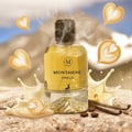 Maison Alhambra Montaigne Vanille by Maison Alhambra 100ml — A clear glass perfume bottle with yellow liquid, a black cap, and a deer silhouette logo, surrounded by latte art hearts, vanilla pods, coffee beans, and milk splashes against a mountainous background. | Maison Alhambra Montaigne Vanille by Maison Alhambra