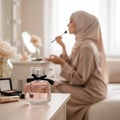 Maison Alhambra My Party Maison Alhambra 100ml EDP Woman — A clear glass perfume bottle with pink liquid and a black bow sits on a wooden table in the foreground, with a woman in a hijab applying makeup in the blurred background. | Maison Alhambra My Party Maison Alhambra 100ml EDP Woman — Botol kaca jernih berisi