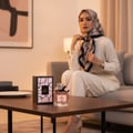 Maison Alhambra My Party Maison Alhambra 100ml EDP Woman — A woman in a hijab and white outfit sits on a sofa, with a perfume bottle and its box on a coffee table in the foreground. | Maison Alhambra My Party Maison Alhambra 100ml EDP Woman — Seorang wanita bertudung dan berpakaian putih duduk di sofa, dengan sebotol