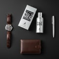 Maison Alhambra No.2 Men by Maison Alhambra 80ml EDP — A flat lay composition featuring a silver perfume bottle and box, a brown leather wallet, a brown leather strap watch, and a silver pen on a dark grey background. | Maison Alhambra No.2 Men by Maison Alhambra 80ml EDP — Komposisi flat lay menampilkan botol dan