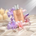 Maison Alhambra Olivia by Maison Alhambra 80ml EDP — A rose gold perfume bottle adorned with wings and the name 'Olivia' is surrounded by purple and pink lilies, with lit candles and pearl-adorned hairpins on satin fabric. | Maison Alhambra Olivia by Maison Alhambra 80ml EDP — Botol minyak wangi berwarna emas ros yang