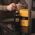 Maison Alhambra Black Origami — Black perfume bottle and yellow box on a reflective black surface, with a person holding a mug in the blurred background. | Maison Alhambra Black Origami — Botol minyak wangi hitam dan kotak kuning di atas permukaan hitam reflektif, dengan seseorang memegang mug di latar belakang kabur.