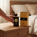 Maison Alhambra Black Origami by Maison Alhambra 100ml EDP — A hand is holding a black perfume bottle with a gold cap on a wooden nightstand next to its gold and black box. | Maison Alhambra Black Origami by Maison Alhambra 100ml EDP — Tangan memegang botol minyak wangi hitam bertutup emas di atas meja sisi kayu