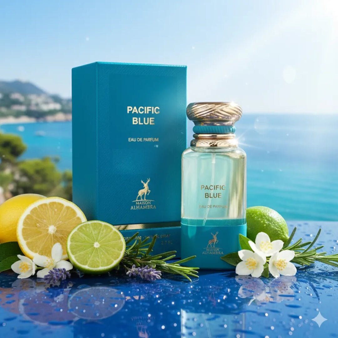Maison Alhambra Pacific Blue by Maison Alhambra 80ml Man — A blue box and a clear bottle of Pacific Blue perfume are displayed on a reflective blue surface with fresh lemons, limes, rosemary, lavender, and jasmine flowers, with a blurred ocean and coastal town in the background. | Maison Alhambra Pacific Blue by