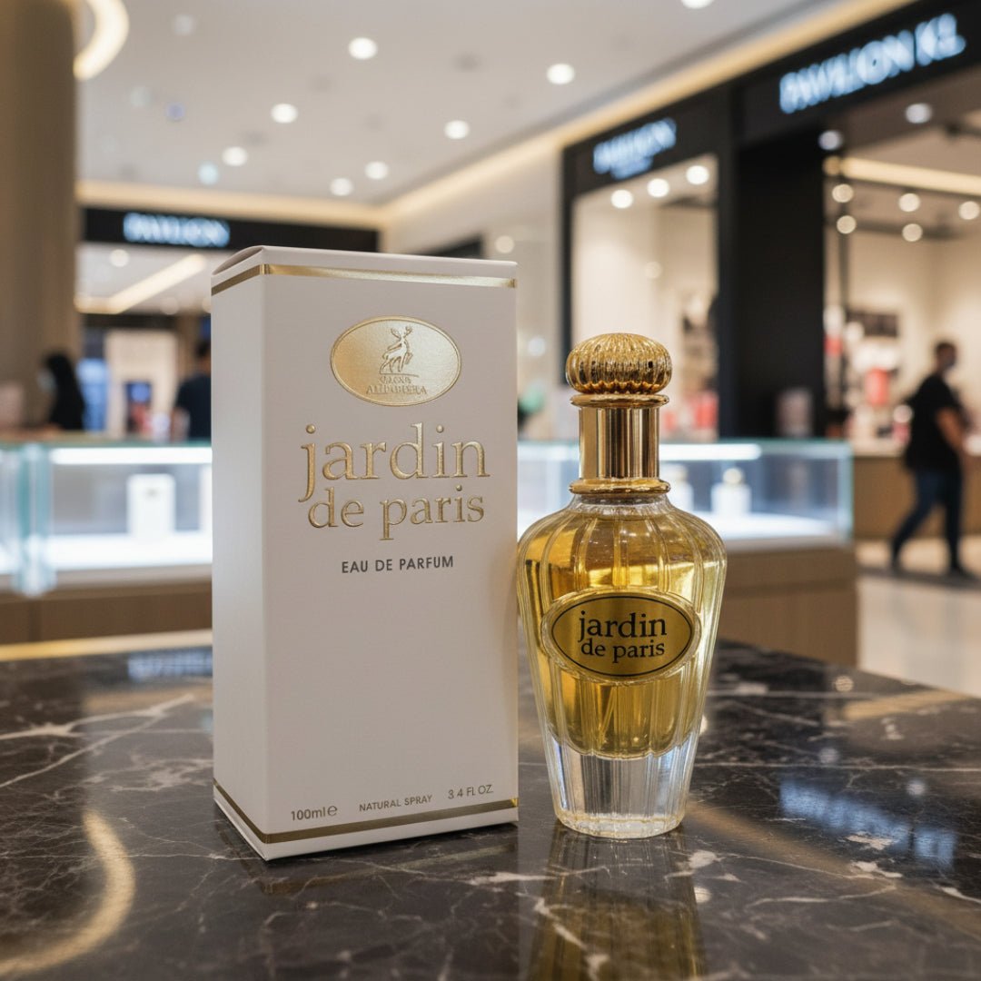 Maison Alhambra Paris Garden 100ml EDP Woman — A white box and a clear glass perfume bottle with a gold cap and gold label are displayed on a marble surface in a shopping mall. | Maison Alhambra Paris Garden 100ml EDP Woman — Peti putih dan botol minyak wangi kaca jernih dengan penutup emas dan label emas dipamerkan