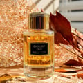 Maison Alhambra Exclusif Oud by Maison Alhambra 100 ml Unisex — A clear glass perfume bottle with golden liquid and a gold cap, set against a textured gold background and dried leaves. | Maison Alhambra Exclusif Oud by Maison Alhambra 100 ml Unisex — Botol minyak wangi kaca jernih dengan cecair keemasan dan penutup