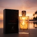 Afnan Toro Pour Homme by Maison Alhambra – A black box and a clear glass perfume bottle with golden liquid are placed on a beach at sunset, with sparkling gold dust in the air. | Afnan Toro Pour Homme by Maison Alhambra – Kotak hitam dan botol minyak wangi kaca jernih berisi cecair keemasan diletakkan di pantai semasa