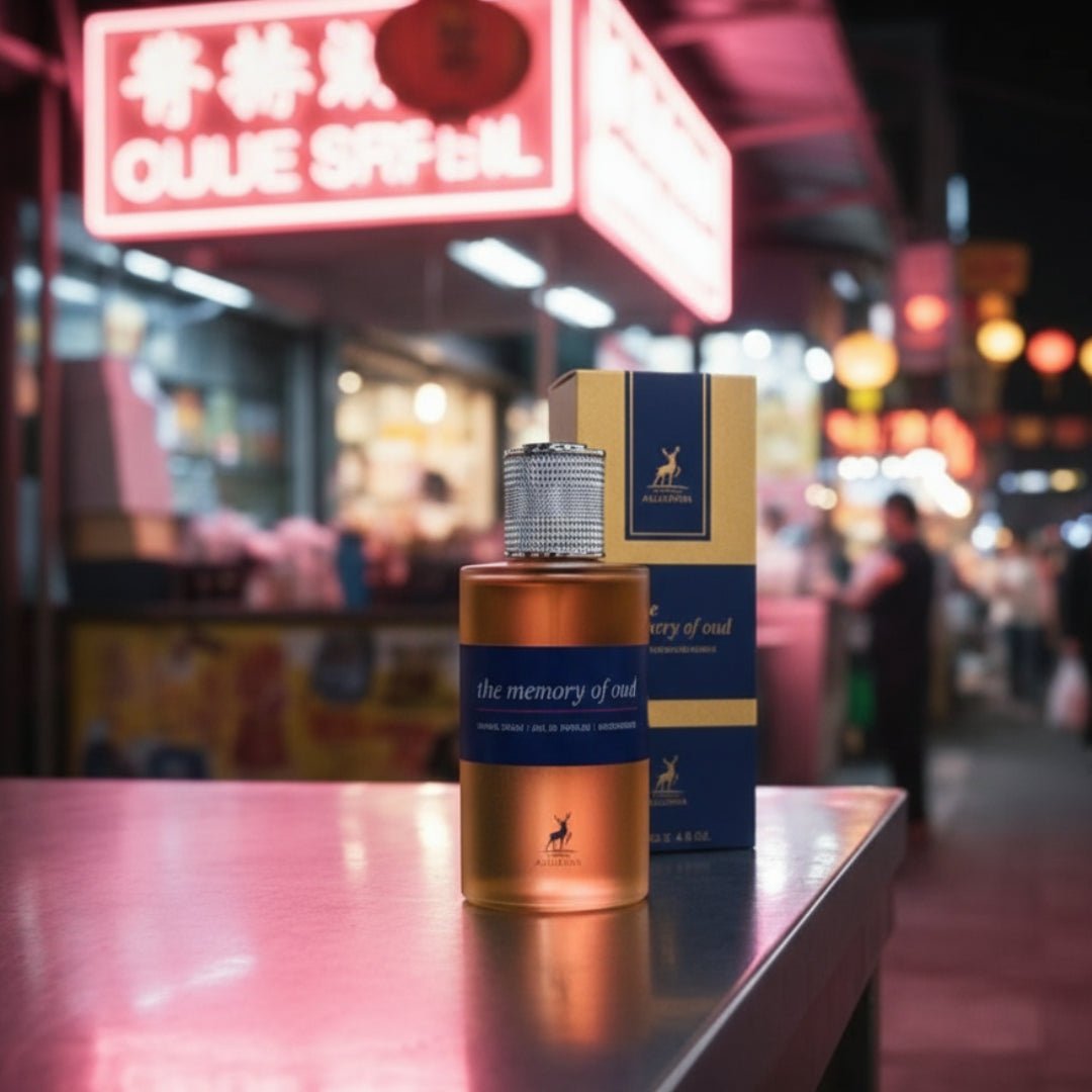 Maison Alhambra The Memory Of Oud EDP 100ml Maison Alhambra — A copper-toned perfume bottle with a textured silver cap sits on a metallic surface, with its blue and gold box behind it. The background is a blurred night market scene with neon signs. | Maison Alhambra The Memory Of Oud EDP 100ml Maison Alhambra — Botol