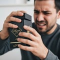 Maison Alhambra Kismet Magic by Maison Alhambra 100ml EDP Unisex — Man with disgusted expression holding a black perfume box with gold lettering, opening the lid. | Lelaki dengan ekspresi jijik memegang kotak minyak wangi hitam dengan tulisan emas, membuka penutupnya.