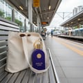 Maison Alhambra Philos Centro 100ml EDP — A purple velvet perfume bottle with a gold cap and ring sits in a canvas tote bag on a train station bench, with sunglasses peeking out of the bag. | Maison Alhambra Philos Centro 100ml EDP — Botol minyak wangi baldu ungu dengan penutup dan cincin emas diletakkan di dalam beg