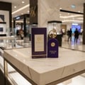 Maison Alhambra Philos Centro — A purple perfume bottle and its matching box sit on a marble surface in a brightly lit store aisle. | Maison Alhambra Philos Centro — Sebotol minyak wangi ungu dan kotak padanannya terletak di atas permukaan marmar di lorong kedai yang terang.