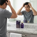 Maison Alhambra Philos Pura by Maison Alhambra 100ml EDP Man — A young man in a grey t-shirt is styling his hair in front of a mirror, with a bottle of Philos Pura and its box on the granite countertop. | Maison Alhambra Philos Pura by Maison Alhambra 100ml EDP Man — Seorang lelaki muda berbaju kelabu sedang