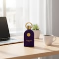Maison Alhambra Philos Pura by Maison Alhambra 100ml EDP Man — A dark purple perfume bottle with a gold ring and emblem sits on a wooden desk next to a laptop and a white mug of coffee, with a potted succulent in the background. | Maison Alhambra Philos Pura by Maison Alhambra 100ml EDP Man — Botol minyak wangi ungu