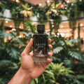 Maison Alhambra Salvo Intense — A hand holds a dark glass perfume bottle with a black cap against a blurred background of lush green plants and warm lights. | Maison Alhambra Salvo Intense — Tangan memegang botol minyak wangi kaca gelap dengan penutup hitam berlatarkan latar belakang kabur tumbuhan hijau subur dan