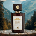 Maison Alhambra Smoked Oud & Vanilla by Maison Alhambra 100ml EDP — A dark brown glass perfume bottle with a wooden cap sits on a rustic wooden table against a blurred background of green mountains and trees. | Maison Alhambra Smoked Oud & Vanilla by Maison Alhambra 100ml EDP — Botol kaca berwarna coklat gelap dengan
