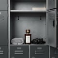 Maison Alhambra Smoked Oud & Vanilla 100ml EDP — A dark glass perfume bottle sits on a shelf in a locker next to a rolled grey towel and black gloves. | Maison Alhambra Smoked Oud & Vanilla 100ml EDP — Sebotol minyak wangi kaca gelap diletakkan di atas rak dalam loker di sebelah tuala kelabu yang digulung dan sarung