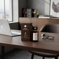 Maison Alhambra Smoked Oud & Vanilla 100ml EDP — A dark brown perfume box and bottle sit on a wooden desk next to a laptop, eyeglasses, and a watch. | Maison Alhambra Smoked Oud & Vanilla 100ml EDP — Kotak dan botol minyak wangi berwarna coklat gelap diletakkan di atas meja kayu bersebelahan dengan komputer riba