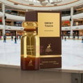 Maison Alhambra Smoky Touch by Maison Alhambra 80ml EDP — A bottle of Smoky Touch perfume with a gold cap and brown liquid sits in front of its brown and gold box, with an ice skating rink and shoppers in the background. | Maison Alhambra Smoky Touch by Maison Alhambra 80ml EDP — Botol minyak wangi Smoky Touch dengan