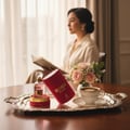 Maison Alhambra So Candid by Maison Alhambra 100 ml EDP — A woman reads a book by a window, with a tray of perfume, roses, and coffee in the foreground. | Maison Alhambra So Candid by Maison Alhambra 100 ml EDP — Seorang wanita membaca buku di tepi tingkap, dengan dulang berisi minyak wangi, mawar, dan kopi di latar