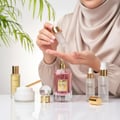 Maison Alhambra So Candid — A woman in a hijab is applying serum from a dropper bottle onto her palm, with the So Candid perfume bottle and other skincare products arranged on a white surface. | Maison Alhambra So Candid — Seorang wanita bertudung sedang menyapu serum dari botol penitis ke tapak tangannya, dengan